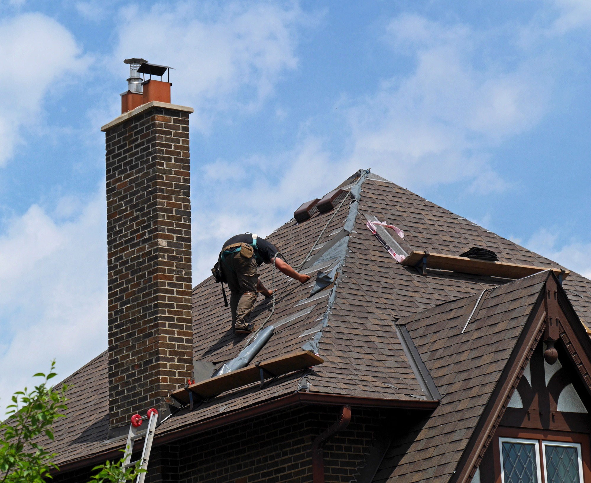 A worker repairs a roof, using tools on sloped shingles near a brick chimney
